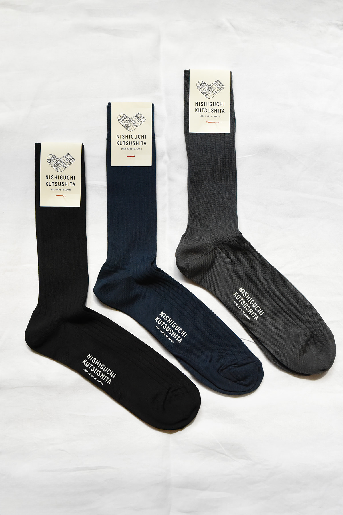 Three pairs of Nishiguchi Kutsushita Silk Cotton Rib Socks in navy, black and charcoal laid flat on a white background with original packaging labels attached.