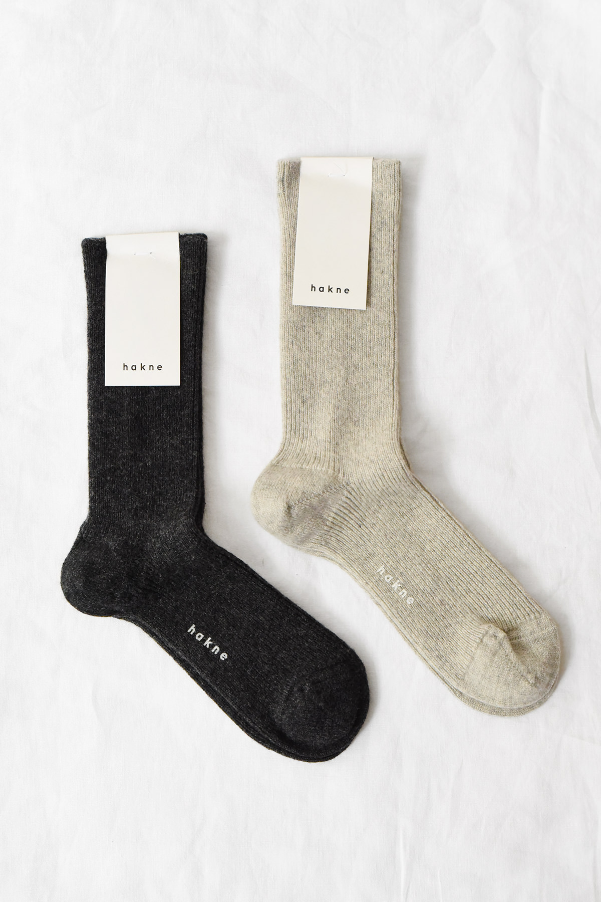 Two pairs of hakne Wool Cashmere Ribbed Socks in dark charcoal and light beige laid flat on a white background with packaging attached.
