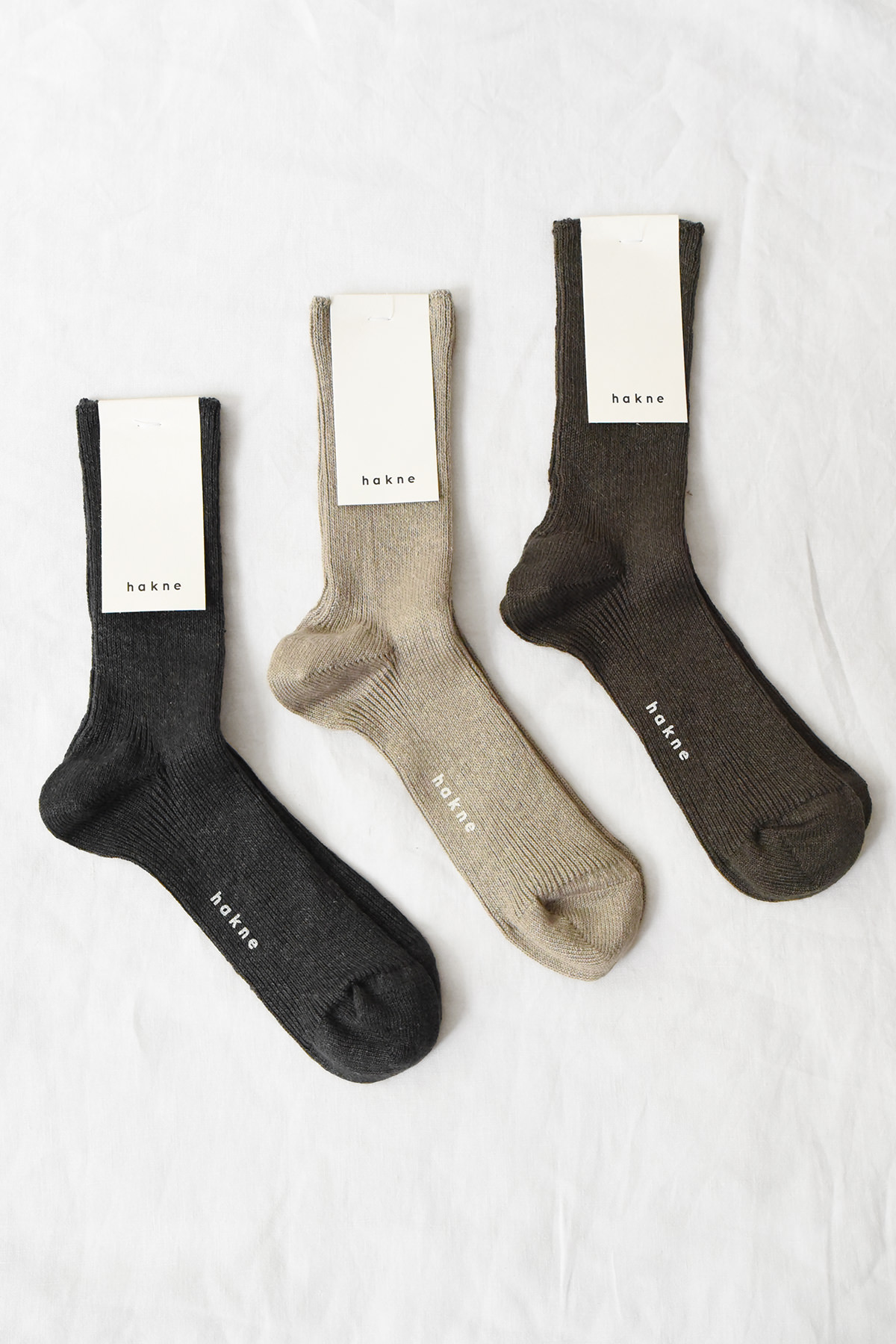 Three pairs of hakne Linen Ribbed Socks in charcoal, mocha brown, and Irish gray laid flat on a white background with packaging attached.