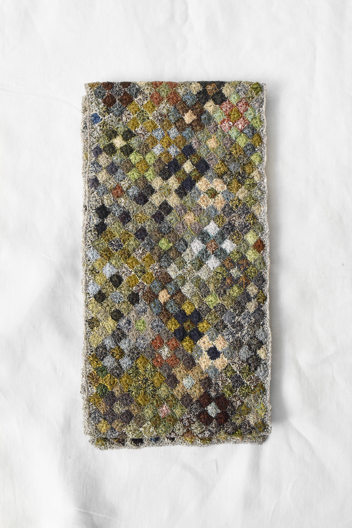 Hand crocheted linen scarf with multicolor patchwork pattern in small square motifs, shown laid flat on a neutral fabric background