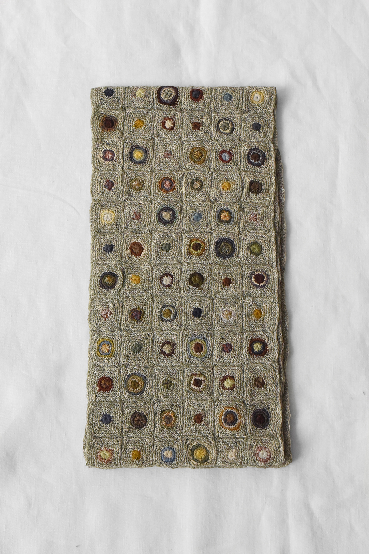 Hand crocheted linen scarf with grid pattern of small square panels featuring circular motifs in varied colors, shown laid flat on a neutral fabric background