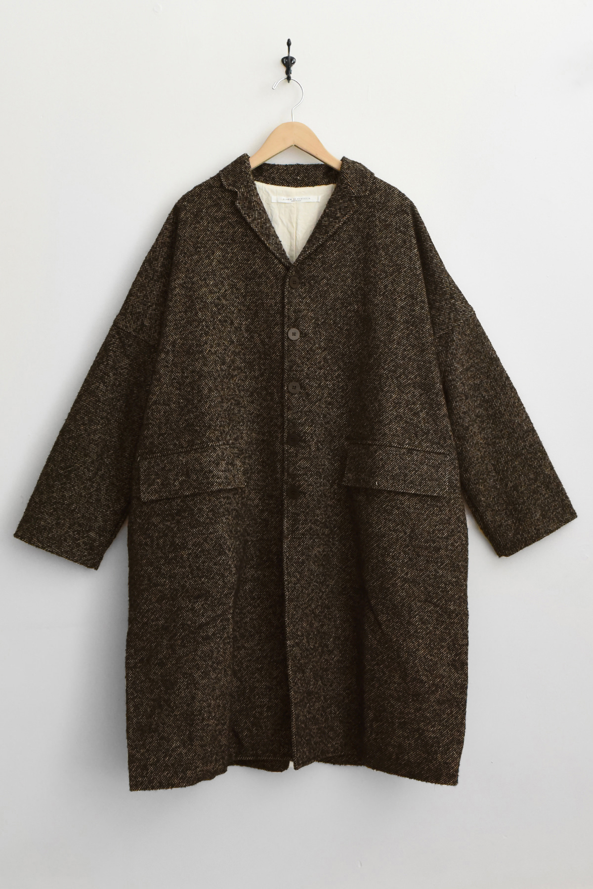 The Album di Famiglia U4558 Loose Coat WN in black, featuring a long, relaxed silhouette with a collar and two large front pockets, is displayed on a wooden hanger against a plain white wall.