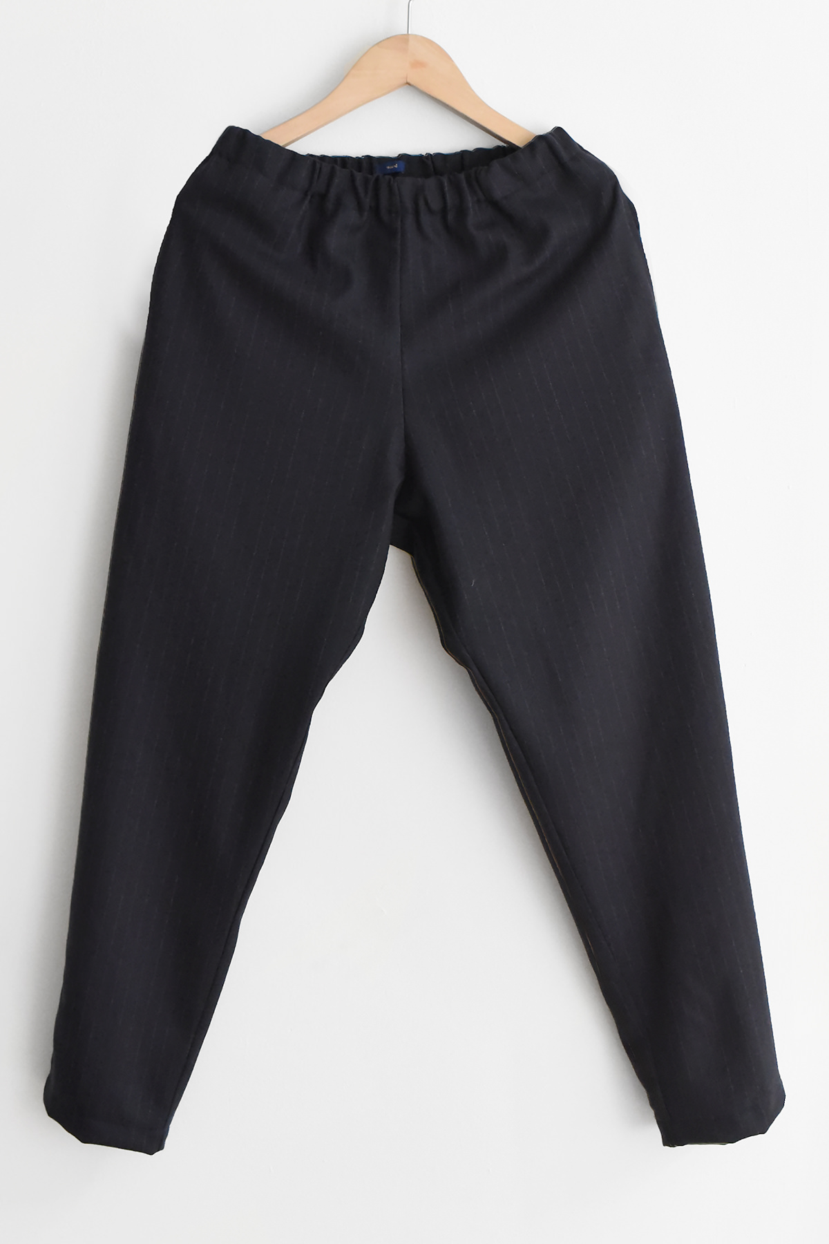 A pair of MAKIE Wool Slim Pants Toni in navy stripe, featuring a straight-leg design and an elastic waistband, is hanging on a wooden hanger against a white background.