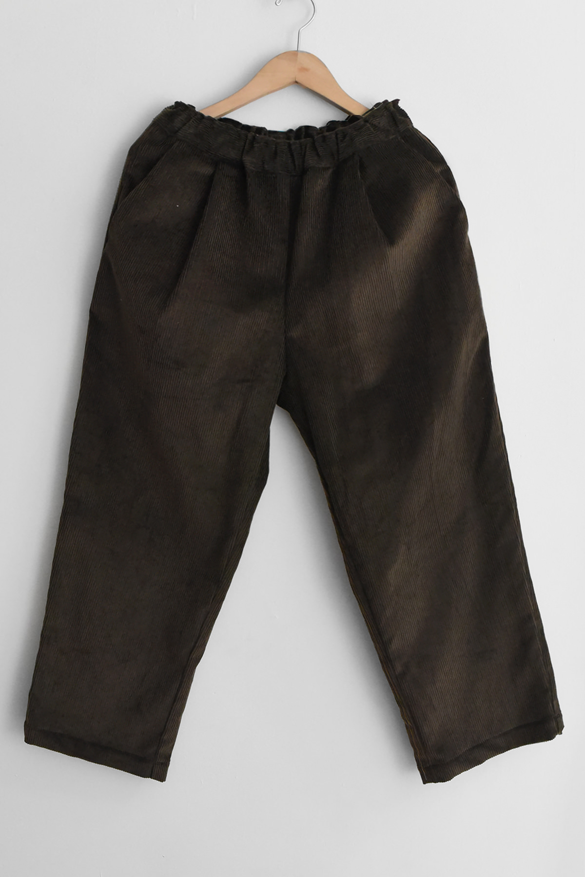 The MAKIE Corduroy Straight Pants Skylar in brown, featuring a loose fit and an elastic waistband, are displayed on a wooden hanger against a white wall.