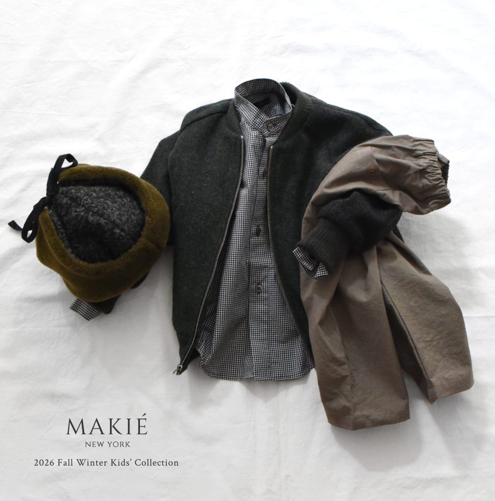 Childrens clothing outfit consisting of a dark zip-up jacket, gingham button-down shirt, brown pants, and a hat with a bow, labeled Makie New York 2026 Fall Winter Kids Collection.