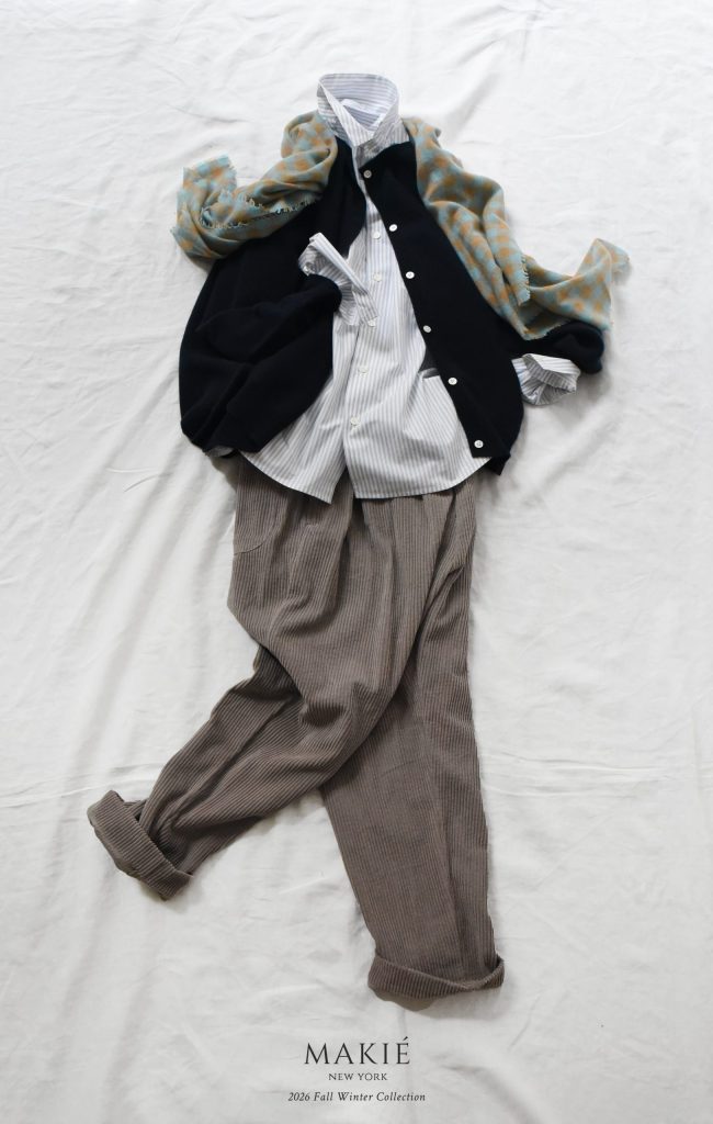 A layered outfit featuring a button-down shirt, dark cardigan, blue and gold checkered scarf, and grey trousers, with Makie New York 2026 Fall Winter Collection text below.