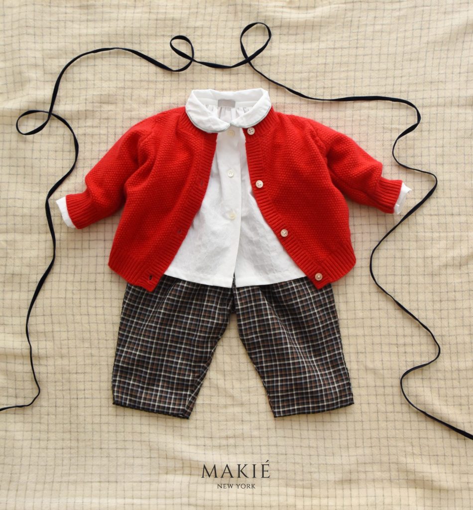 Childs outfit consisting of a red button-down cardigan, white collared shirt, and plaid trousers, laid flat on a grid-patterned fabric with black ribbon trim.