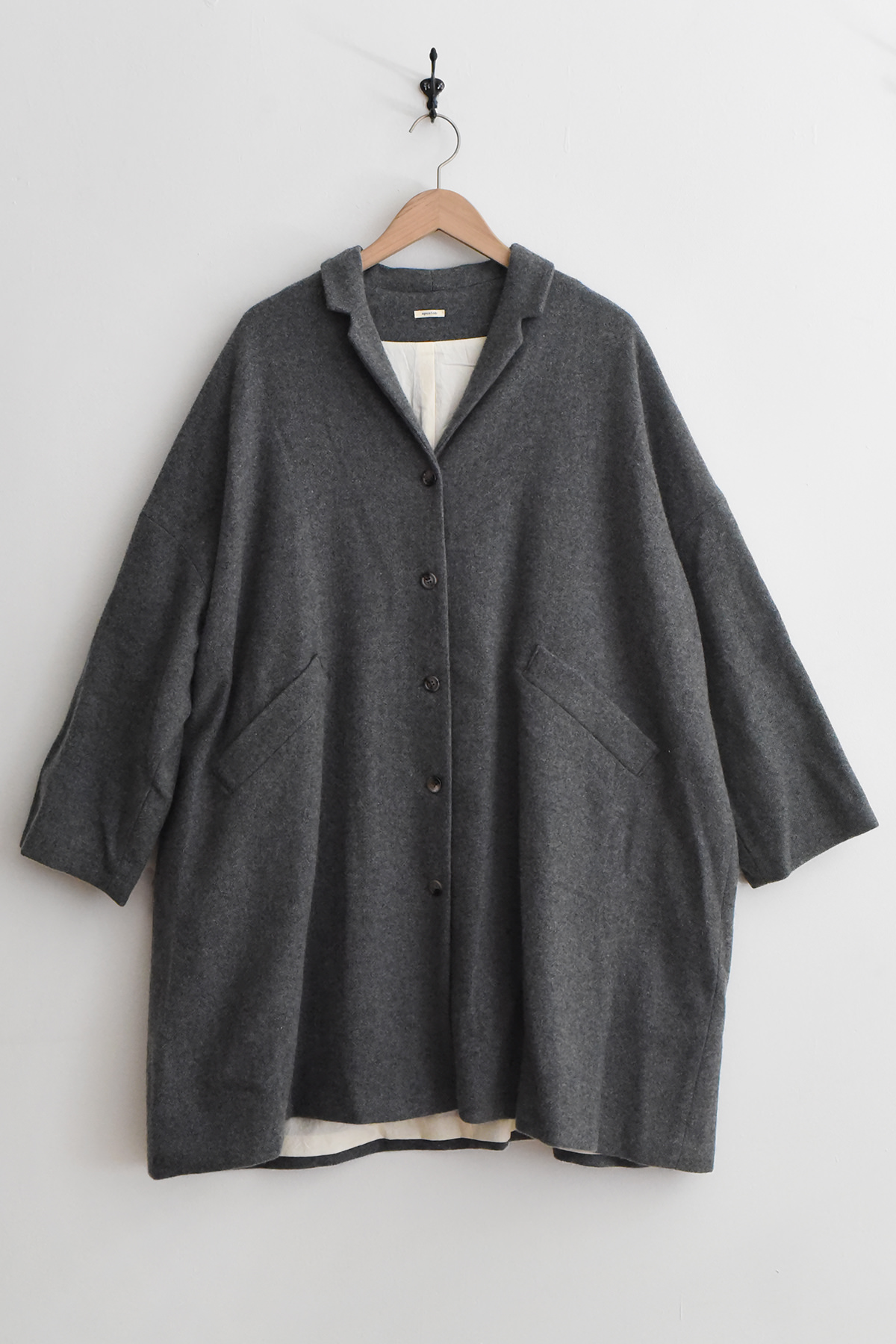 Apuntob (a.b) P2087 Coat - Cement in an oversized grey design with a notched collar, button front, and angled front pockets, hanging on a wooden hanger against a white wall.