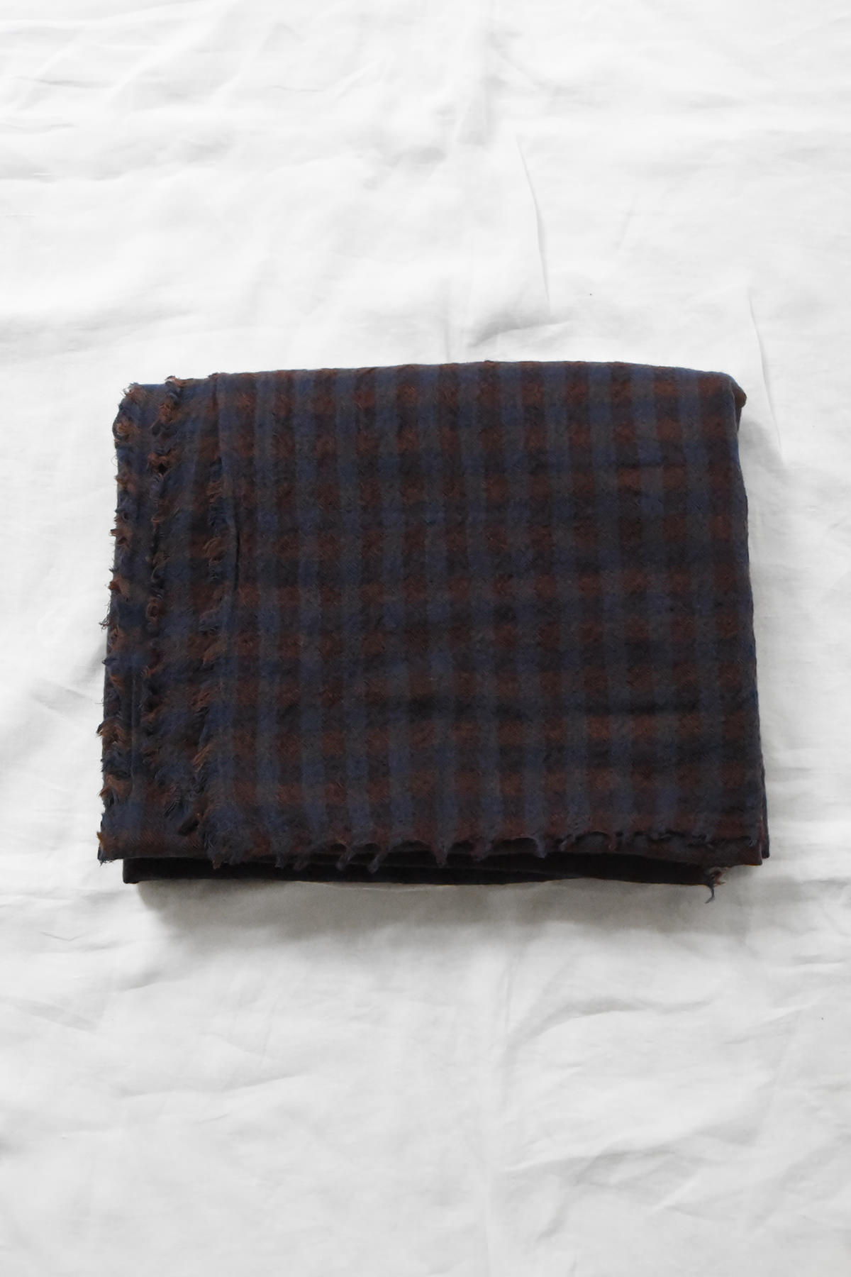 Manuelle Guibal, 7146 Wool Scarf YSH - Noctis in a folded rectangular shape with a dark blue and brown checkered pattern and raw, fringed edges.