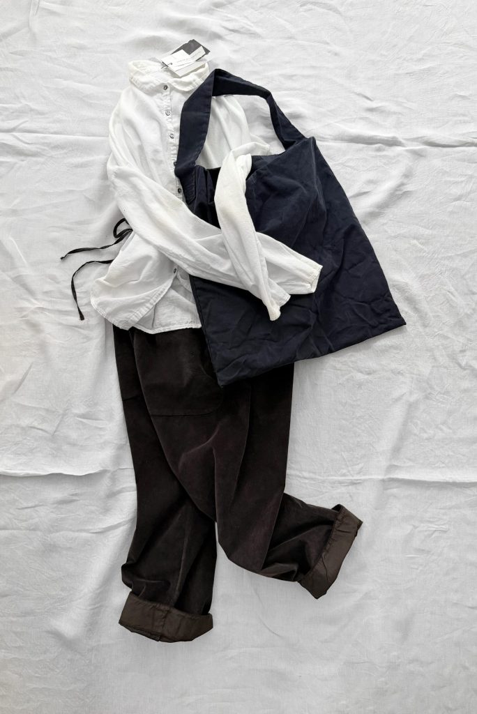 A white button-down shirt, dark trousers, and a black tote bag laid out on a white fabric surface.