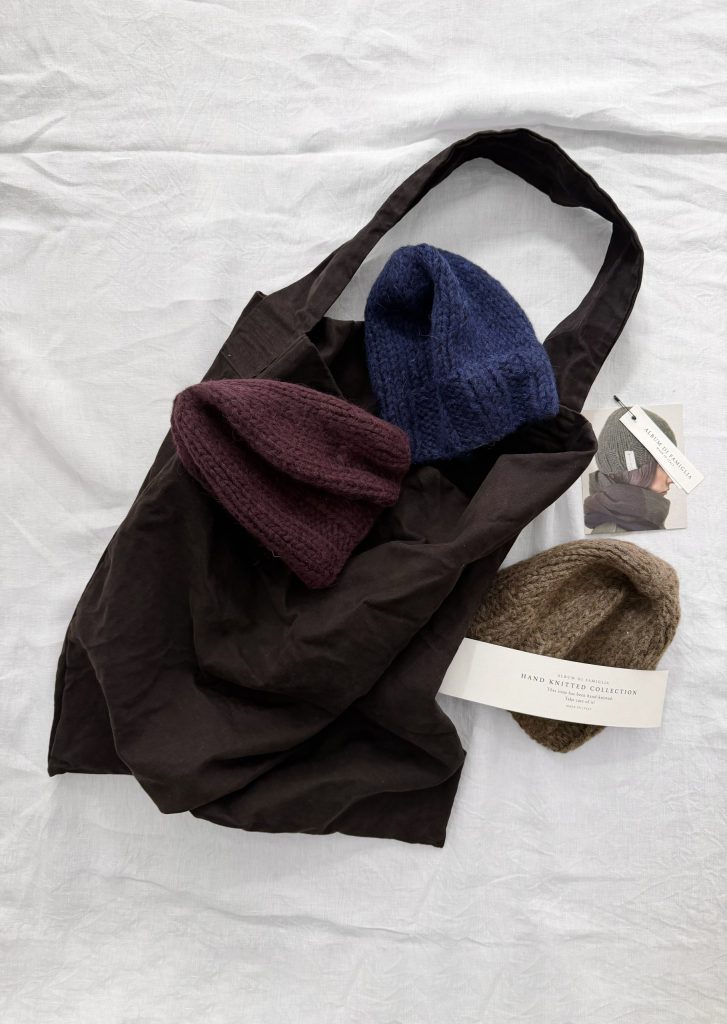 A dark brown fabric tote bag containing three knit beanies in burgundy, navy, and brown, with two product tags featuring the Album Di Famiglia hand-knitted collection branding.